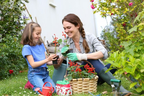 A woman and a young girl are gardening together in a lush outdoor garden, surrounded by vibrant green foliage, flowering plants, and a well-maintained lawn. The woman, wearing green gardening gloves and casual attire, is smiling as she holds a small potted plant, while the girl, dressed in a blue T-shirt and denim shorts, is carefully handling the same plant. In front of them, there are gardening tools and supplies, including a woven basket, a watering can, and plant pots placed on the grass. The background features dense shrubbery, flowering bushes, and tall plants, creating an inviting and lively garden environment typical of residential outdoor spaces in Finsbury Park. The scene captures a moment of shared outdoor activity, emphasizing garden care and planting, relevant to professional gardening services offered by Landscapers Finsbury Park. The lighting indicates a clear, sunny day, enhancing natural colours and textures of the garden elements.