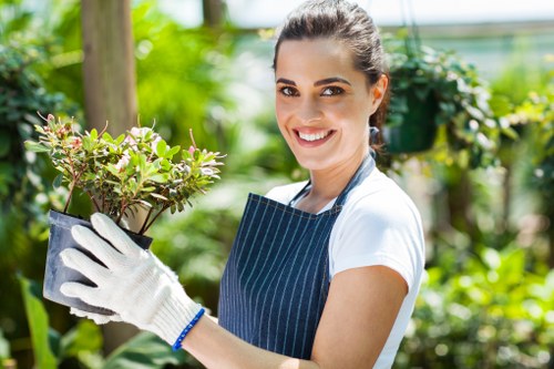 A young woman with dark hair tied back, smiling warmly while wearing a white t-shirt and a dark blue striped apron, holds a potted shrub with small, green leaves and pinkish flowers in a lush garden setting. The garden features a variety of green foliage, including hedges, trees, and flower beds, with a wooden post visible in the background. The scene is well-lit with natural daylight, highlighting the vibrant greens and the woman's cheerful expression. This outdoor space exemplifies well-maintained landscaping suitable for gardening and lawn care services, reflective of residential or community gardens in Finsbury Park, London, with a focus on sustainable and eco-friendly practices as indicated on the 'Recycling and Sustainability' local webpage of Landscapers Finsbury Park.
