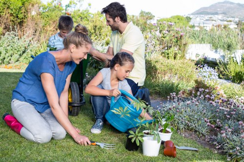 A family gardening together in a well-maintained garden, with a young girl placing a plant into the soil while seated on the grass, assisted by an adult woman. In the background, a man and another child are seen, engaging with the outdoors. The garden features a lush, green lawn with a variety of flowering plants and shrubs bordering the space. There are gardening tools and containers, including a watering can and small pots, placed on the grass beside the family. The scene is set outdoors during daylight with clear weather, showcasing a tidy and inviting landscaped yard that includes gravel pathways and wooden decking. This natural, vibrant environment reflects professional gardening and landscaping efforts typical of services offered by Landscapers Finsbury Park, focused on outdoor maintenance and garden enhancement in the London area.
