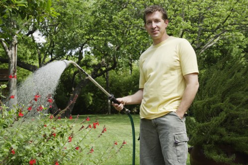 A man standing in a well-maintained front garden during daytime, holding a garden hose and watering the plants. The garden features a neatly trimmed lawn with lush green grass, bordered by flowering shrubs with red blossoms and various bushes. In the background, there are tall trees with dense foliage providing shade, and a wooden fence partially visible behind the greenery. The scene is natural and bright, suggesting pleasant weather, and the lawn appears healthy and vibrant. This outdoor space exemplifies typical garden features, aligned with landscaping and gardening services offered by companies like Landscapers Finsbury Park in the local area around N4 postcode, contributing to a tidy, thriving garden environment.