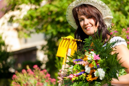 A woman in a garden during daytime, holding a yellow gardening trowel and a vibrant bouquet of various flowers and green foliage. She is wearing a wide-brimmed straw hat and a sleeveless top, with a smiling expression. The garden features lush greenery, including a hedge or shrub in the background, with a mix of flowering plants and foliage in the foreground. The outdoor space appears well-maintained, with natural lighting highlighting the rich greens and bright colors of the flowers, typical of a landscaped garden in Finsbury Park. This scene aligns with gardening and outdoor maintenance services provided by Landscapers Finsbury Park, set within an inviting residential garden environment.