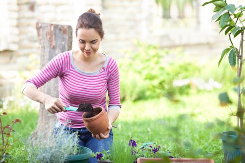 A woman with dark hair tied back, wearing a pink and white striped long-sleeve top, is gardening in a backyard garden during daylight hours. She is holding a small terracotta pot in her right hand and using a trowel in her left hand to work with dark, moist soil inside the pot. The garden features a lush, green lawn in the foreground, with various potted plants and flower beds surrounding her. In the background, there is a stone wall, a tree trunk, and leafy shrubs that add natural texture to the outdoor space. The scene is well-lit by natural sunlight, suggesting a clear day. This setting is indicative of professional gardening or outdoor maintenance activities commonly carried out by landscapers and garden specialists in Finsbury Park, with the focus on planting and garden care.