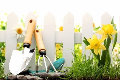 A landscaped garden scene featuring a white picket fence in the background with bright yellow daffodils and green foliage in the foreground. In front of the fence, there is a patch of well-maintained, lush green grass and a small flower bed bordered by soil. Gardening tools, including a trowel and a small garden fork with wooden handles, rest on the grass, indicating ongoing or recent garden maintenance. The setting appears to be outdoors on a clear, sunny day, with natural light illuminating the vibrant colours of the flowers and greenery. This garden reflects elements typical of a residential outdoor space in the Finsbury Park area, suitable for professional gardening services like those offered by Landscapers Finsbury Park. The scene emphasizes the importance of lawn care, flower bed maintenance, and outdoor aesthetics, aligning with professional landscaping and gardening upkeep in the local area.