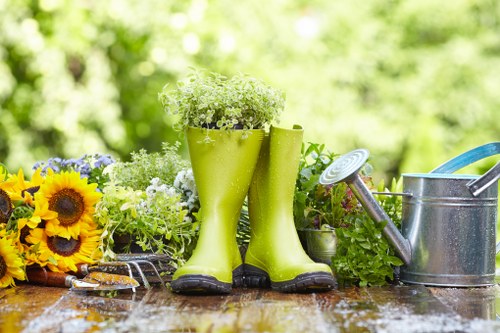 The image features a garden scene with a wooden outdoor table displaying gardening tools and decorative items. In the foreground, there are bright yellow sunflower-themed decorations and a pair of lime green rubber boots filled with white flowering plants, suggesting recent planting or decoration. To the right, a metal watering can with a blue handle and a silver spout is positioned beside potted green plants, including a small fern and other leafy foliage. The background shows a lush, green garden with dense foliage and trees, illuminated by natural sunlight, creating a warm and inviting outdoor environment. The wooden surface of the table appears slightly wet, indicating recent watering or rain, and the overall scene reflects a well-maintained garden suitable for gardening and outdoor plant care activities. The scene aligns with landscaping and gardening services offered by Landscapers Finsbury Park in the N4 postcode area, emphasizing plant nurturing, lawn maintenance, and outdoor garden enhancements.