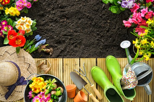 The image shows a well-maintained garden area with a focus on a small wooden decking section bordered by a lush, green lawn. Alongside the decking, there are various gardening tools and accessories including bright green rubber gardening boots, a small metallic watering can, a pair of gardening gloves with a floral pattern, and a trowel placed nearby. In the top left corner, a wicker sun hat with a decorative band rests alongside a black pot filled with colorful flowers such as pink, yellow, and purple blooms. The background features a neat, dark soil bed bordered by a row of flowering plants, including pink, yellow, and purple blossoms, creating a vibrant floral border. The overall scene suggests an outdoor gardening space ready for planting or maintenance, with natural lighting indicating a calm, sunny day, suitable for outdoor gardening or landscaping work by a provider like Landscapers Finsbury Park in the N4 postal area.