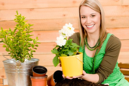 The image depicts a cheerful woman with blonde hair smiling as she holds a yellow flower pot containing a lush white flowering plant, set against a warm wooden backdrop in a garden setting. In front of her are various gardening containers, including a large metallic pot with a green shrub, a black plant pot, and an orange flower pot, indicating she is engaged in gardening activities. The foreground features a well-maintained, vibrant green lawn with neatly trimmed grass, complemented by a garden bed or border located just behind her, which is edged with soil or mulch and contains a few small plants or shrubs. The environment appears to be outdoors on a bright, mild day, with soft natural light highlighting the healthy, densely packed grass and the greenery surrounding her. This outdoor scene is characteristic of a backyard or front garden designed for leisure and gardening, aligning with the services offered by landscapersfinsburypark.co.uk for lawn care and garden maintenance in Finsbury Park or nearby areas, reflecting a well-tended outdoor space suitable for residential gardening activities.