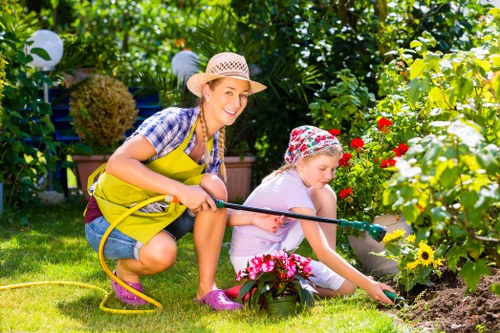 In a vibrant, well-maintained front garden, a woman and a young girl are engaged in gardening activities under bright sunlight, with lush green trees and neatly trimmed hedges providing a natural backdrop. The woman, wearing a straw hat, yellow apron, and casual clothing, smiles as she crouches beside the girl, who is wearing a floral hat and light-colored outfit. They are tending to flower beds filled with colorful blooms, including red, pink, and yellow flowers, with the girl gently planting or watering the plants. The garden features a lush, green lawn, bordered by flower beds with rich, dark soil and mulched edges, and paved pathways unseen but implied by the spatial arrangement. In the background, decorative garden lanterns hang among the trees, adding a charming touch to the outdoor space. The scene captures a peaceful moment of outdoor gardening, illustrating the importance of garden maintenance and green space care, typical of services provided by Landscapers Finsbury Park in the local area around N4 postcode, with natural tones and textures emphasizing a healthy, well-kept garden environment suitable for both recreation and aesthetic enhancement.