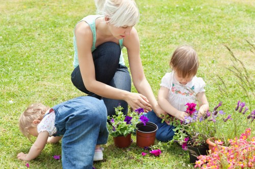 A woman with short platinum blonde hair, wearing a light blue sleeveless top and dark blue jeans, is kneeling on a well-maintained green lawn in a garden during daytime. She is engaged in planting or arranging potted plants, with her hands near a small, empty terracotta pot. To her right, a young girl with light brown hair, dressed in a white top with a floral pattern, is sitting on the grass, holding a pink flower and examining a flowering plant in a black pot. To her left, another young girl with curly blonde hair, wearing a denim dress, is crawling on the grass, reaching towards the ground. The garden features lush, dense grass with some flowers in bloom, including purple and pink blossoms positioned in pots and beds. In the background, there are shrubbery and trees, suggesting a tidy and landscaped outdoor space. The scene is naturally lit, indicating clear weather, and the setting reflects a typical residential outdoor area suitable for gardening and family activities in Finsbury Park or nearby areas. This image supports gardening and outdoor maintenance services, such as planting, lawn care, and garden landscaping offered by Landscapers Finsbury Park.