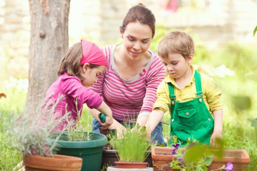 A woman with dark hair tied back, wearing a pink and white striped long-sleeve top, is engaging in gardening activities with a young girl and boy in a lush, green garden. The girl, dressed in a pink jacket and pink headband, is working with a small gardening tool in a large turquoise planter filled with green plants. The boy, wearing a yellow shirt with green overalls, stands nearby observing attentively. The garden features a well-maintained lawn area, a sturdy tree trunk visible in the background, and several flower pots and planters with various plants and flowers, suggesting a space designed for outdoor planting and nurturing. The scene is set outdoors during daylight, with natural sunlight illuminating the vibrant colors of the flowers, plants, and garden surfaces, offering a peaceful outdoor environment that supports gardening services such as planting, maintenance, or general garden care in the Finsbury Park area. The environment emphasizes community engagement with gardening activities, highlighting the natural textures of soil, foliage, and garden tools on the scene.