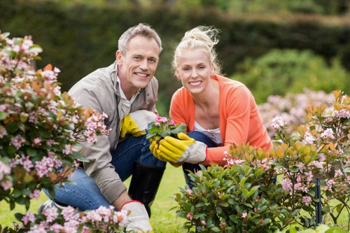 A man and a woman are kneeling in a well-maintained garden with lush green grass and flowering shrubs, smiling at the camera while engaging in gardening activities. They are dressed casually, wearing gardening gloves, and surrounded by vibrant pink and white blossoms, with a backdrop of neatly trimmed hedges and trees. The garden features a combination of flowering plants and shrubs, creating a colorful and inviting outdoor space typical of residential gardens in the Finsbury Park area. The scene is illuminated by natural daylight, suggesting pleasant weather suitable for outdoor maintenance and gardening work carried out by professionals like Landscapers Finsbury Park. The overall setting showcases a tidy, landscaped yard with a focus on flowering plant beds and managed lawn areas, reflecting services related to lawn care, plant maintenance, and garden enhancement.
